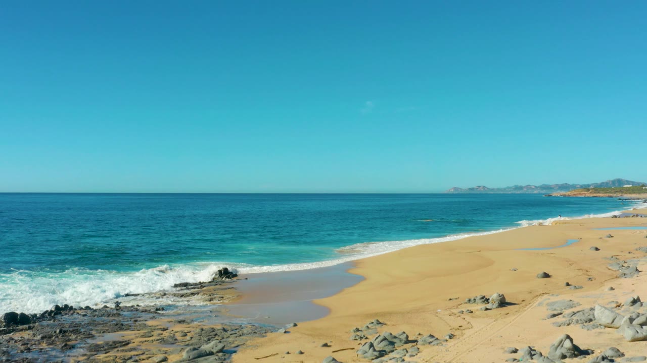 Laguna Beach Los Cabos shoreline with white waves curling onto sand, scattered beach rocks, deep blue water offshore, clear sky, and no visible people.