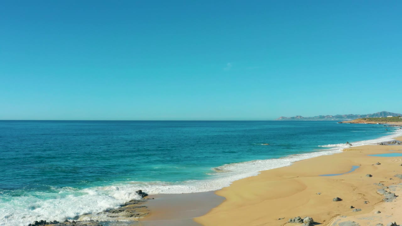 Elevated coastal view at Laguna Beach Los Cabos showing open blue ocean, a pale sand beach at right, white shore break, distant hills, and no visible people.