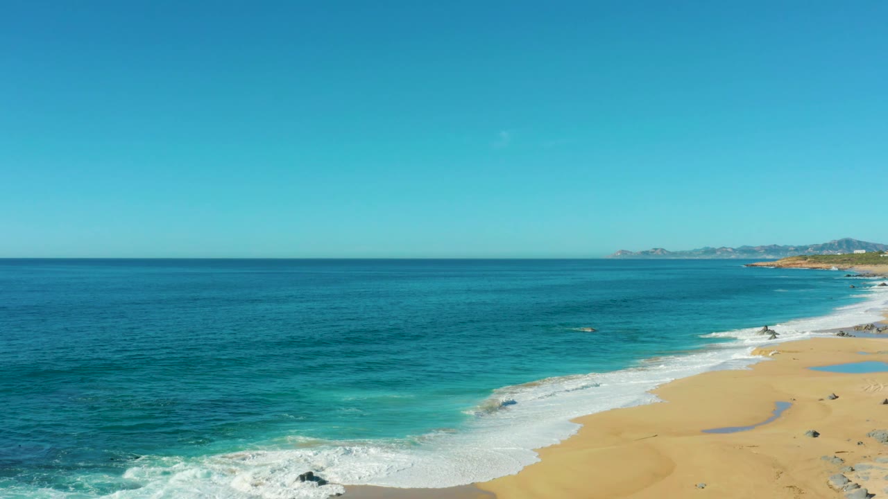 Laguna Beach Los Cabos coastline with broad blue ocean filling most of the frame, sandy beach along the lower right, white surf, distant land, and no visible people.