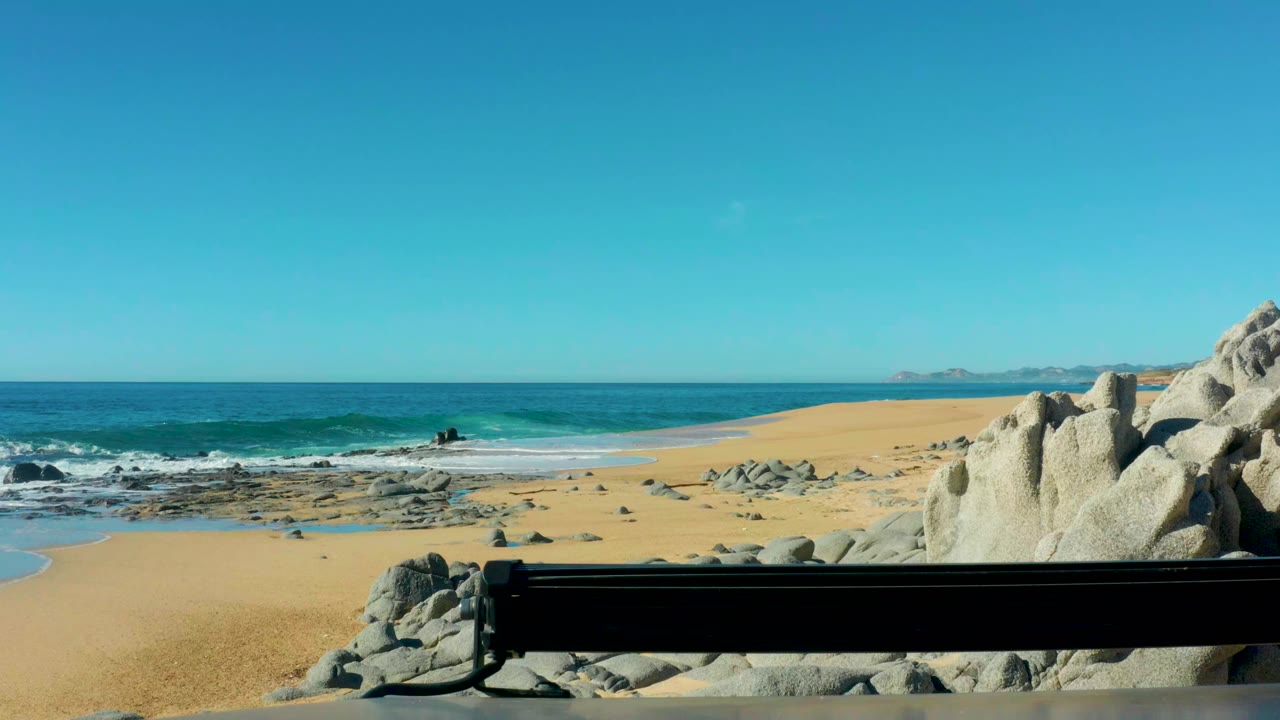 Wide beach view with surf, rocks, clear sky, and a dark bench in the foreground.