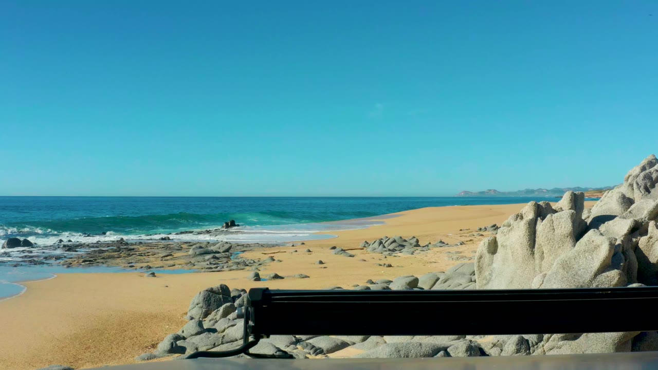 Laguna Beach Los Cabos beach scene with a small wave forming near the shore, a black bench across the lower frame, right-side rocks, and no visible people.