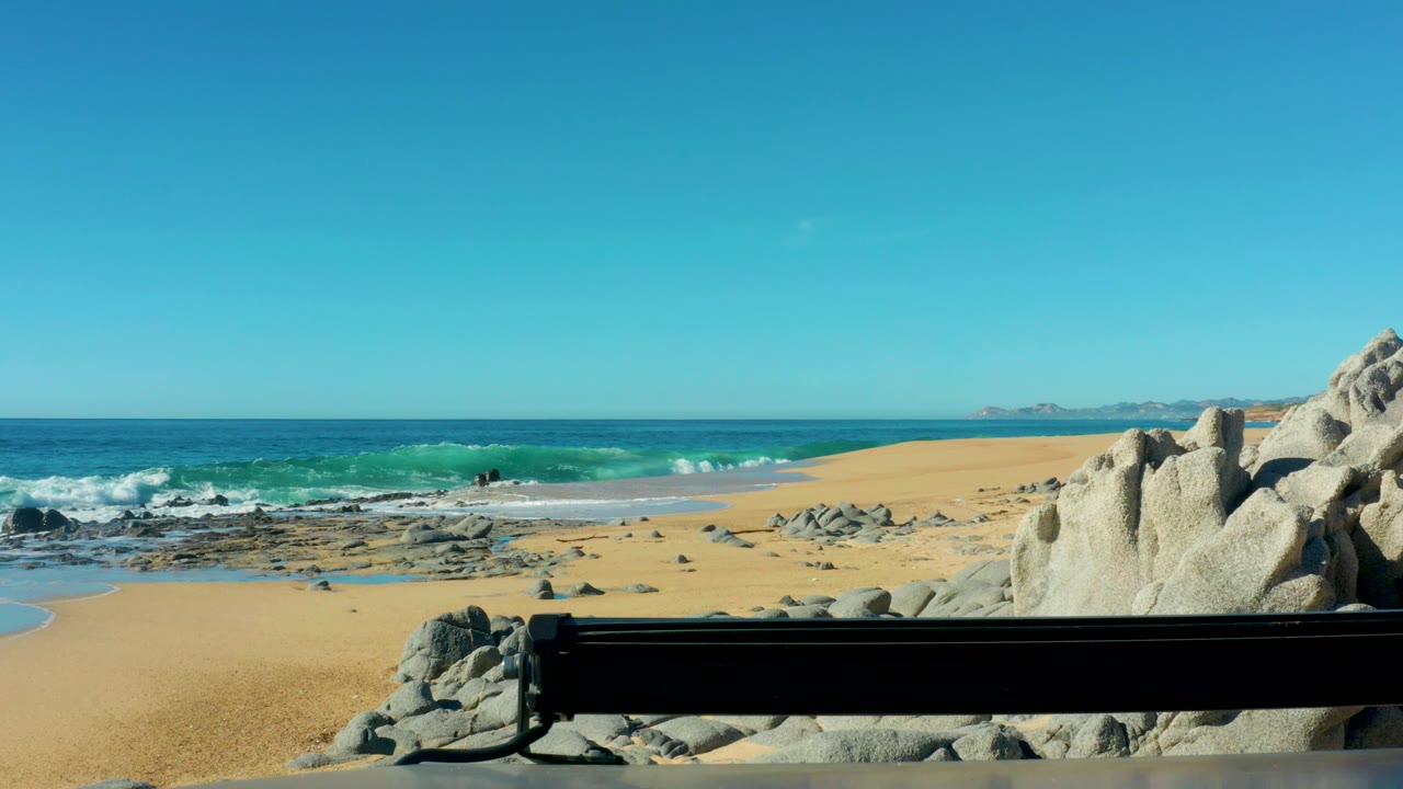 Laguna Beach Los Cabos beach view with foam rolling over the near shoreline, scattered dark rocks in the surf, pale boulders at right, and no visible people.