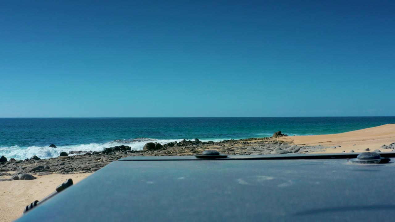 Laguna Beach Los Cabos view with tan sand, dark rocks, turquoise ocean, and a dark foreground surface across the lower frame, no visible people.