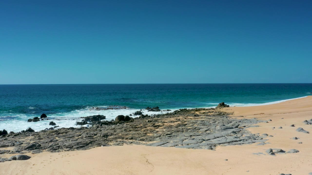 Laguna Beach Los Cabos coast with a broad sand foreground, low dark rocks, white waves rolling in, and no visible people.