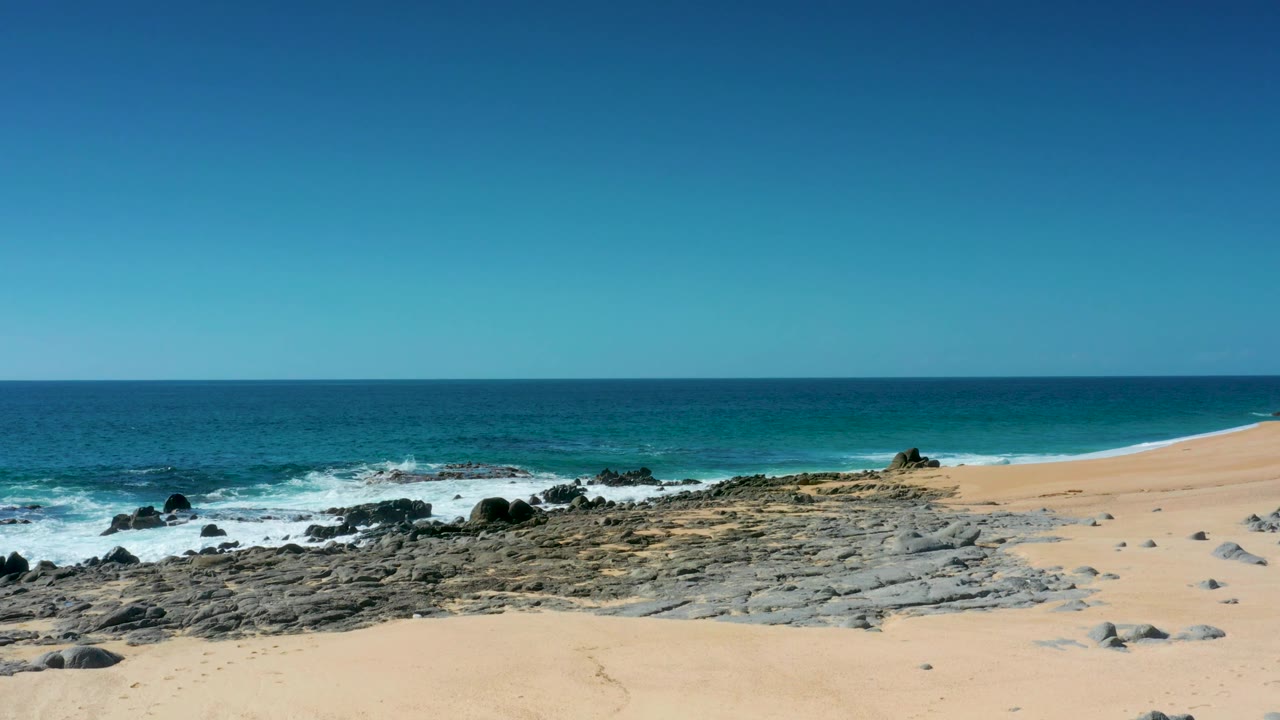 A wide view of Laguna Beach Los Cabos shows open sand, scattered rocks near the surf, blue-green water, and no visible people.