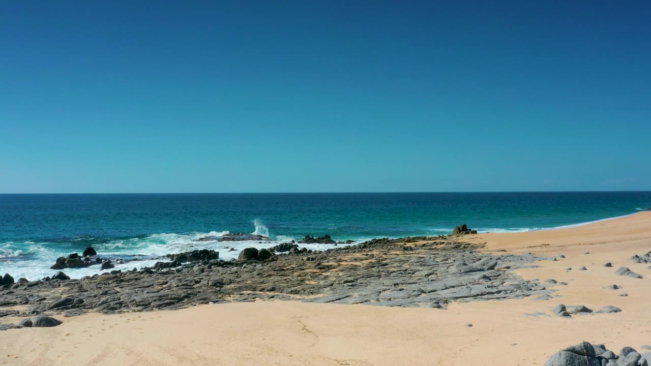 Laguna Beach Los Cabos beach scene with textured sand, dark rocks, breaking whitewater, a straight ocean horizon, and no visible people.