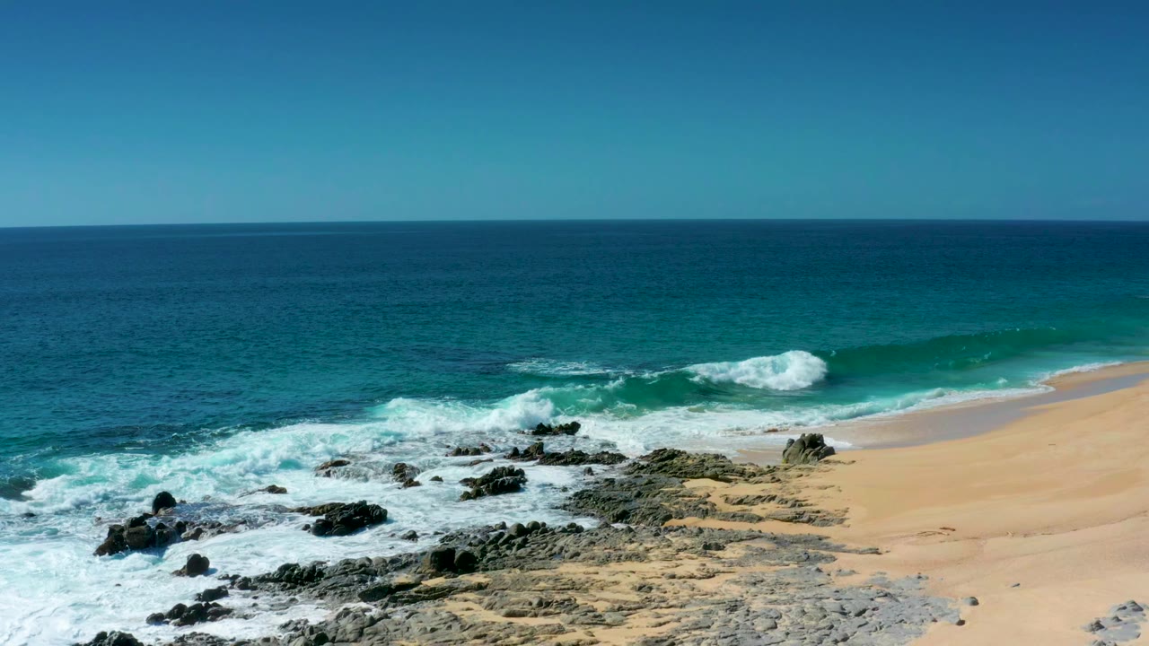 At Laguna Beach Los Cabos, white foam spreads over the black rocks while a clean sandy slope runs along the right side with no visible people.