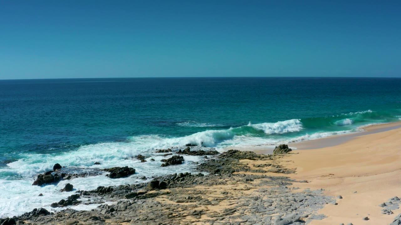 At Laguna Beach Los Cabos, a wave breaks farther along the shoreline, with rough rocks in the foreground and empty sand on the right with no visible people.