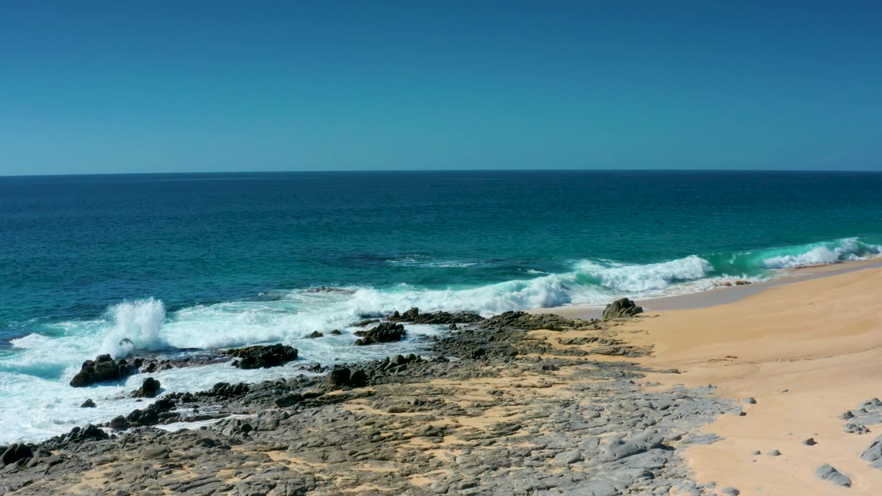 At Laguna Beach Los Cabos, surf splashes up at the left edge of the rock shelf, with turquoise water beyond and no visible people on the sand.