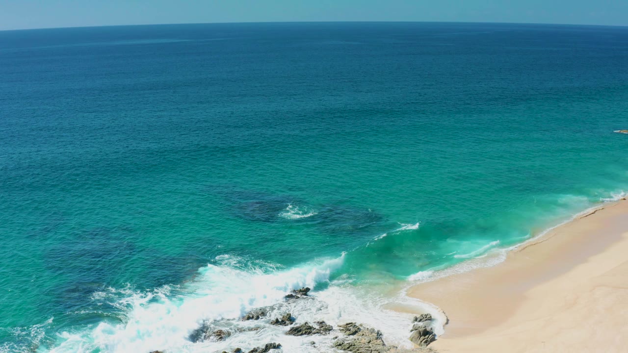Wide aerial view of Laguna Beach Los Cabos with turquoise water, white surf hitting dark shoreline rocks, pale sand at the right edge, and no visible people.