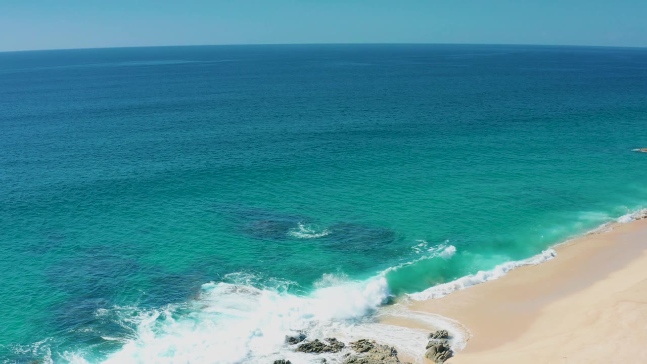 Aerial shoreline view in Laguna Beach Los Cabos with bright white wave wash curling over dark rocks, clear turquoise shallows, open sea beyond, pale sand to the right, and no visible people.
