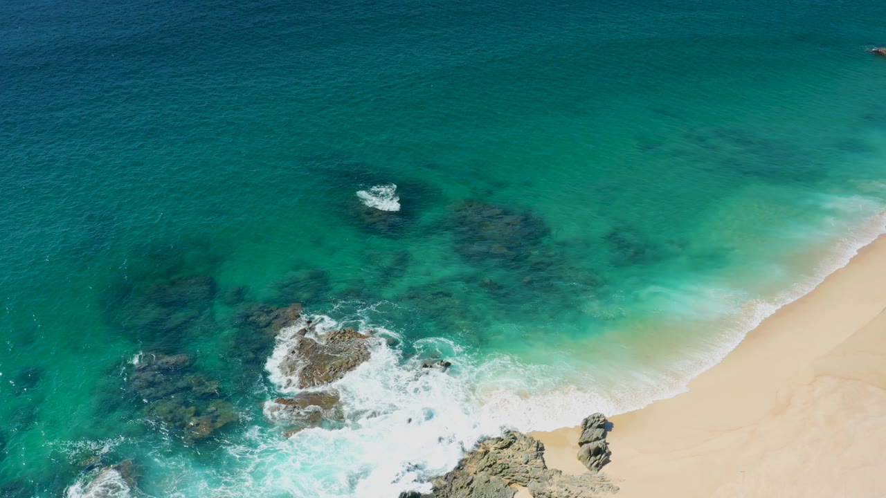 Aerial view in Laguna Beach Los Cabos with foam spreading around the central rock outcrop and the sandy beach curving along the right edge, with no visible people.