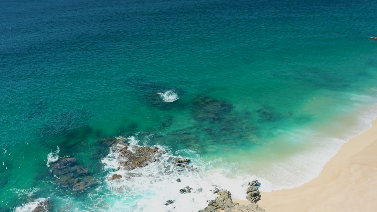 Aerial frame from Laguna Beach Los Cabos with more open water above the rocks, foamy surf at the lower left, and sand along the right side, with no visible people.