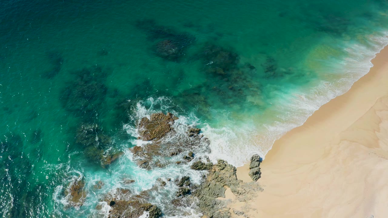 Aerial view of turquoise surf breaking over rocks beside sand