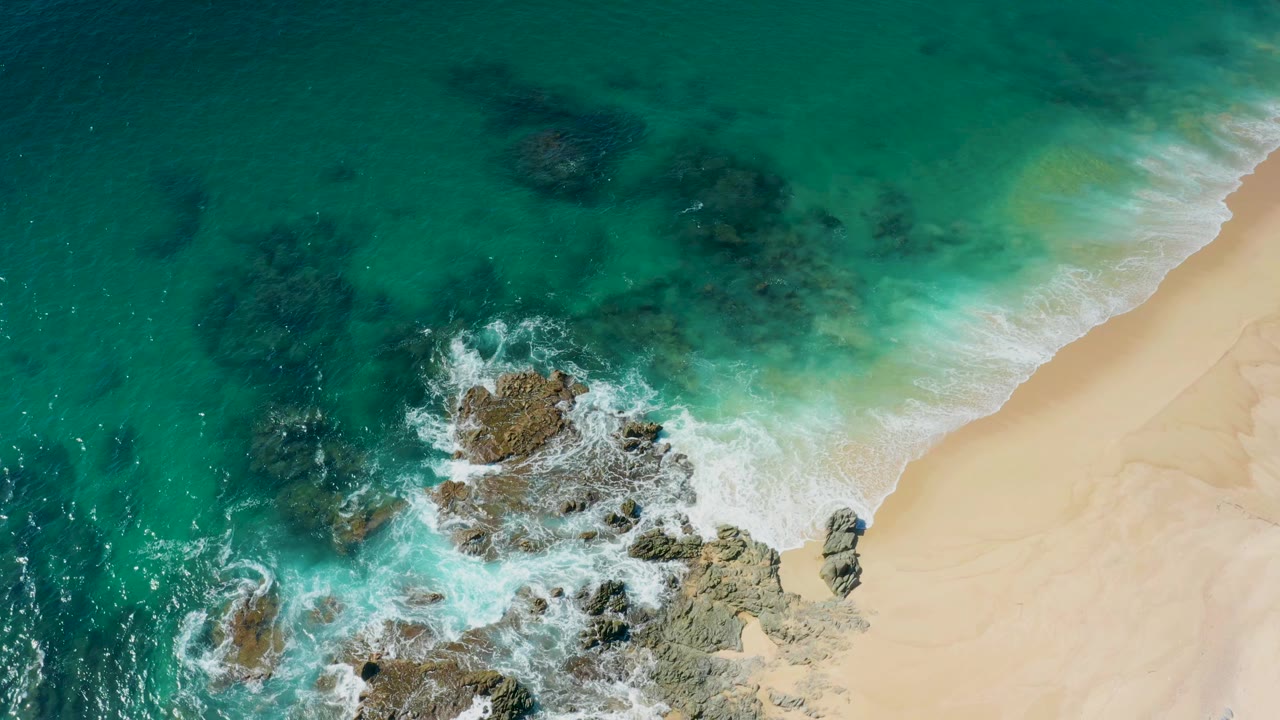 Overhead view at Laguna Beach Los Cabos with white surf spreading around central rocks, pale sand along the right edge, and no visible people.