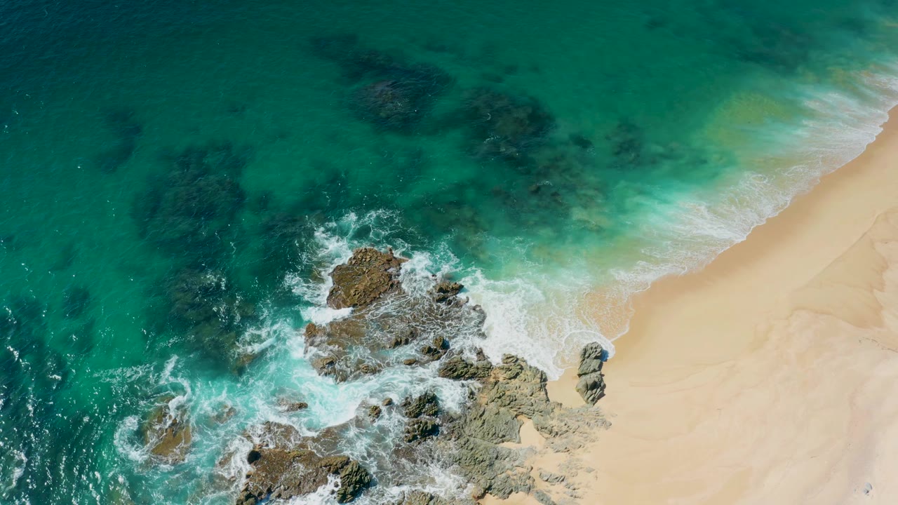 Laguna Beach Los Cabos shoreline seen from above as foamy water wraps a larger rock cluster and the sandy beach curves through the right side, with no visible people.