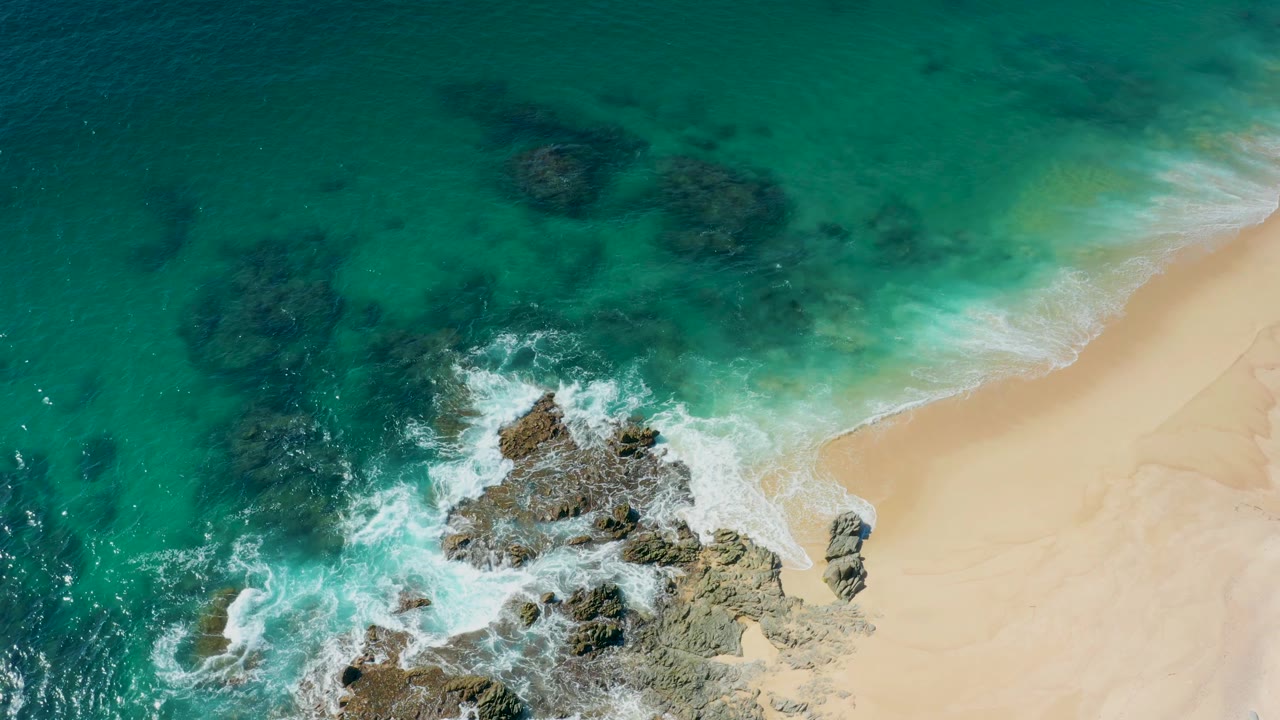 Overhead Laguna Beach Los Cabos frame showing dark submerged rocks, a foamy break near exposed stone, and a clean stretch of sand with no visible people.