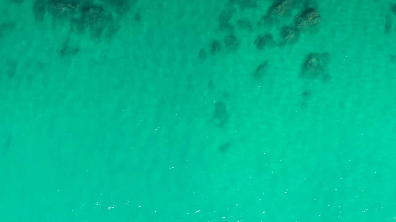 Clear green-blue ocean water at Laguna Beach Los Cabos shows scattered dark rocks below the surface, faint sand streaks, and no visible people.