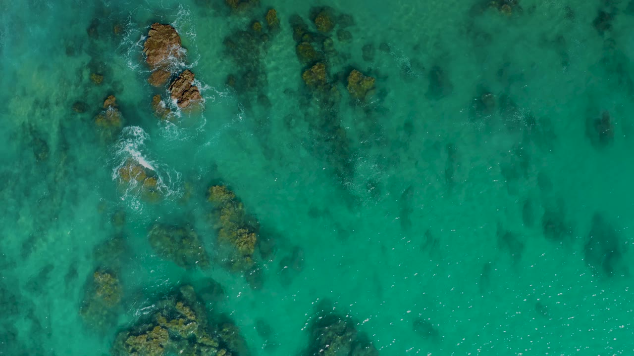Overhead view of clear turquoise water at Laguna Beach Los Cabos with submerged rocks near the left side, small white foam around the upper rocks, and no visible people.