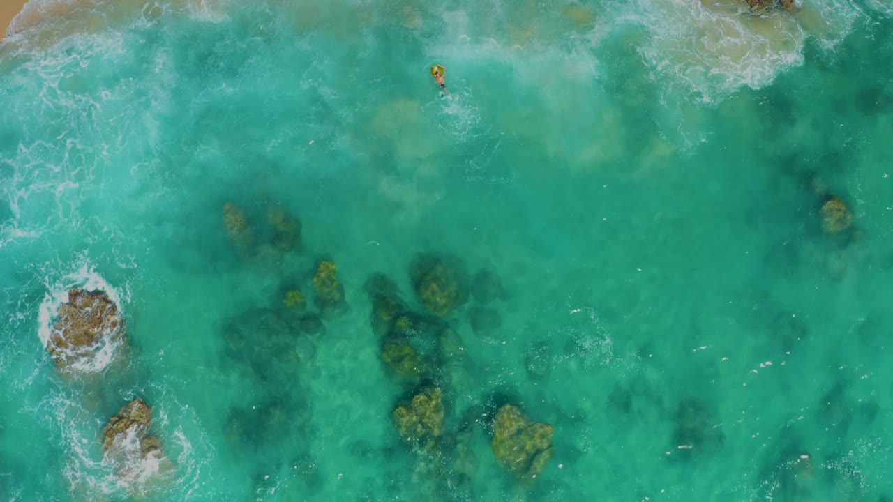 Drone view of Laguna Beach Los Cabos water where a larger rock breaks the surface near the lower left, smaller submerged rocks sit to the right, and no visible people appear.