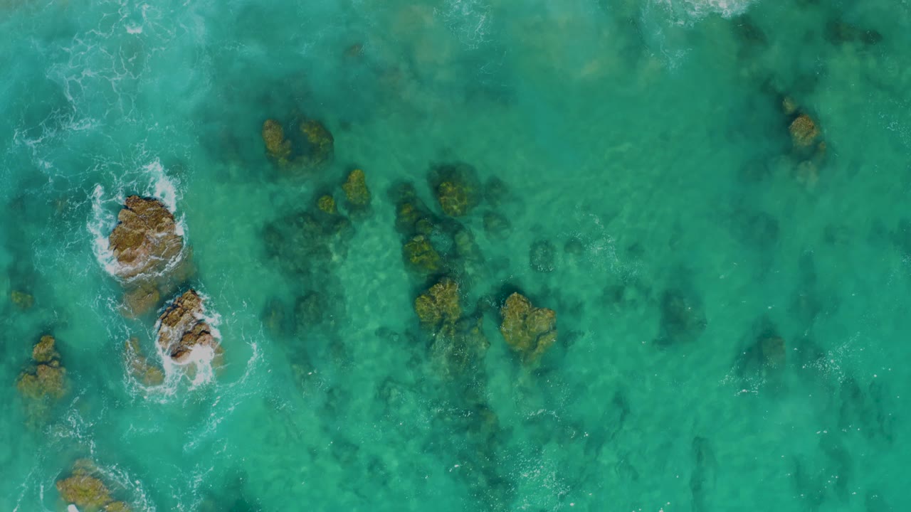 Top-down view at Laguna Beach Los Cabos with a foamy rock cluster left of center, small rocks around it, clear green-blue water, and no visible people.