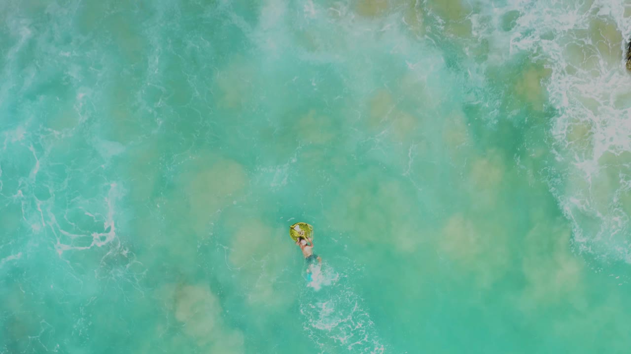 Overhead view at Laguna Beach Los Cabos showing turquoise water, white surf texture, and a small visible person on a yellow float below scattered shoreline foam.