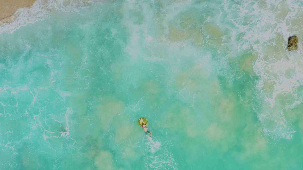 Overhead Laguna Beach Los Cabos view with a visible person on a yellow float near the lower right, breaking whitewater above, and a strip of sand at the top edge.