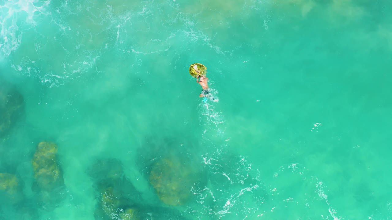 Overhead frame from Laguna Beach Los Cabos showing a person near the upper center holding a yellow inflatable ring in clear turquoise water with darker underwater rocks below.
