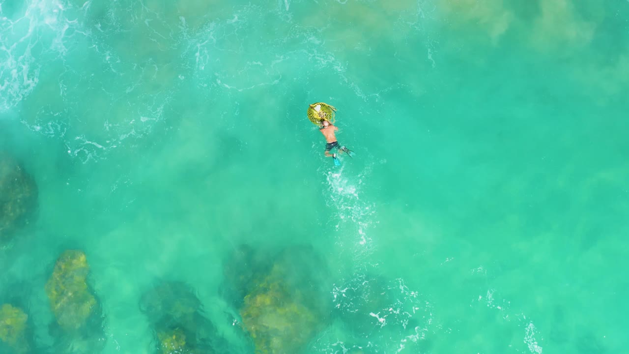 At 0:02, the overhead view keeps the person small against a wide field of turquoise water, with the yellow ring near the upper right and submerged rock patches lower in frame.
