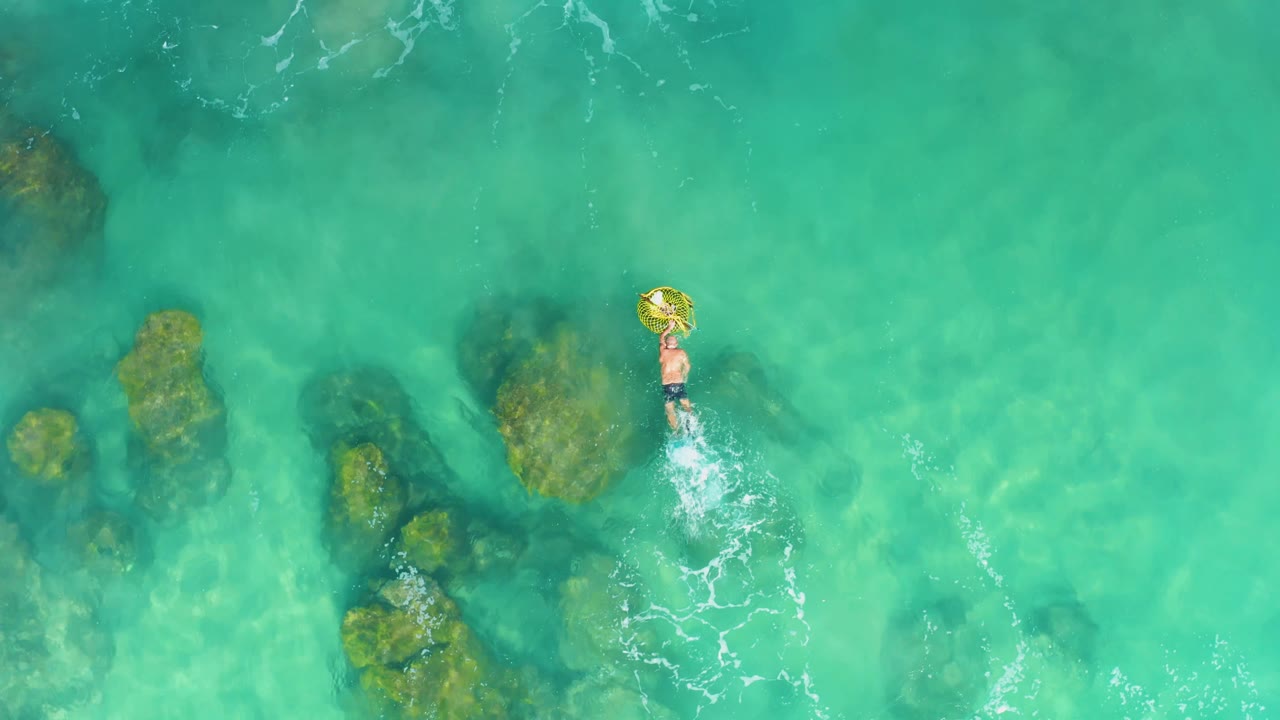 A top-down drone view shows the person with the yellow ring slightly right of center at Laguna Beach Los Cabos, with scattered submerged rocks and small white wakes behind the swimmer.