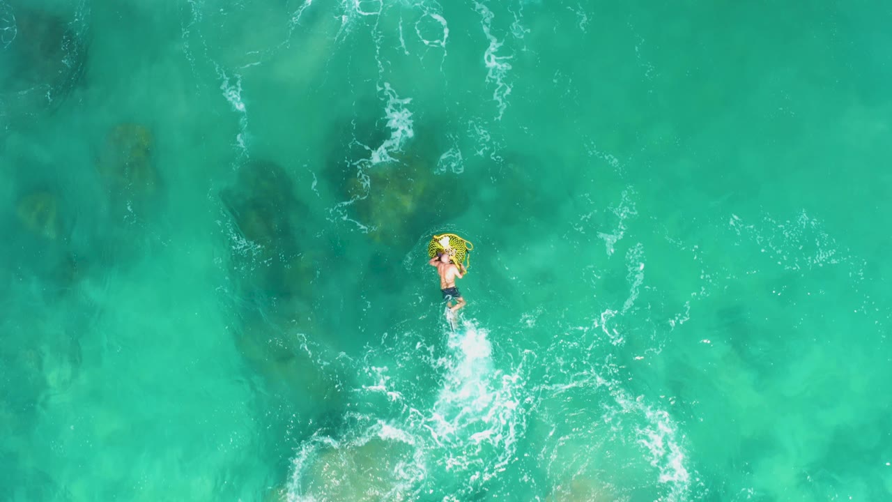 Overhead frame from Laguna Beach Los Cabos showing a person on a yellow inflatable ring near the upper center, with white splash trails in clear turquoise water.