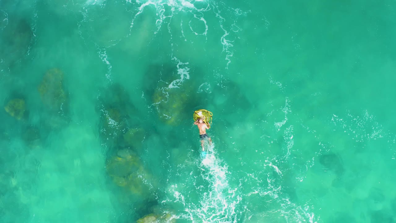 Overhead Laguna Beach Los Cabos view with the person on the yellow inflatable ring farther right in the frame and scattered submerged rocks visible beneath the clear water.