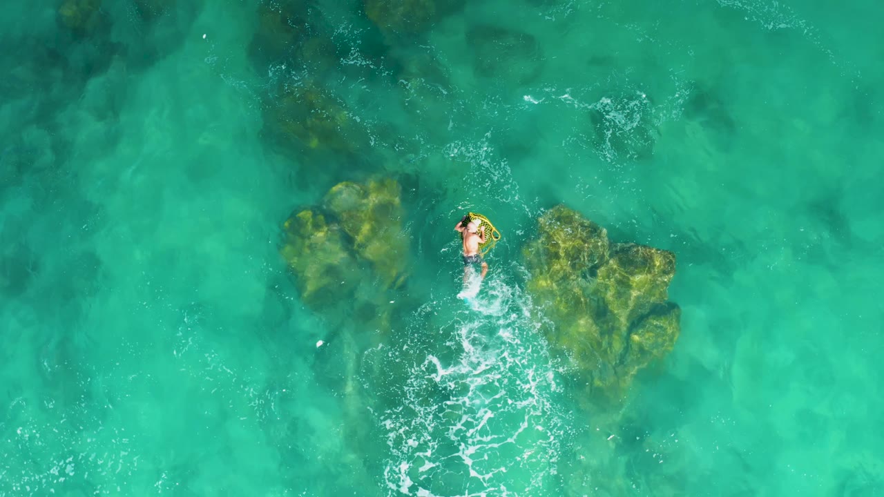 At Laguna Beach Los Cabos, the person swims just above the main submerged rocks, with pale foam and clear turquoise water around the body.