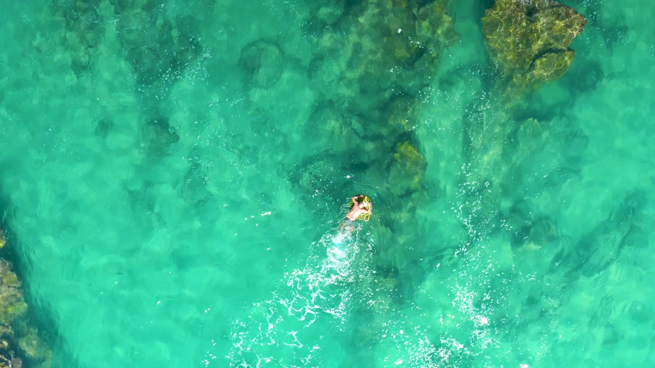 Overhead frame from Laguna Beach Los Cabos showing a swimmer near the lower center moving through clear turquoise water with submerged rocks above.