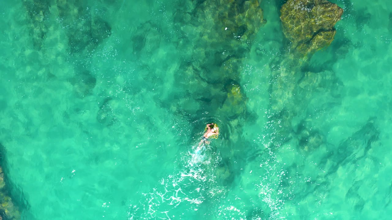 Overhead frame from Laguna Beach Los Cabos with the swimmer near the right side, white churn behind them, and dark rock patches visible through the water.