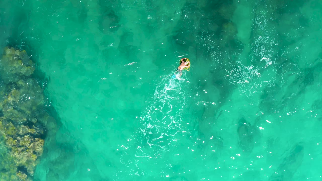 Swimmer crossing clear turquoise ocean water with rocks below.