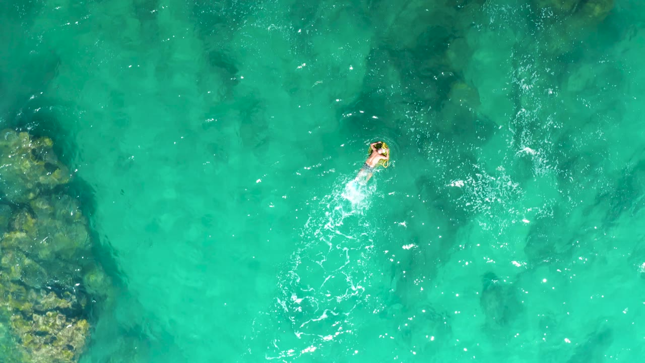 Top-down Laguna Beach Los Cabos frame with the swimmer near the upper right area, white surface ripples trailing behind, and dark rock patches visible under the turquoise water.