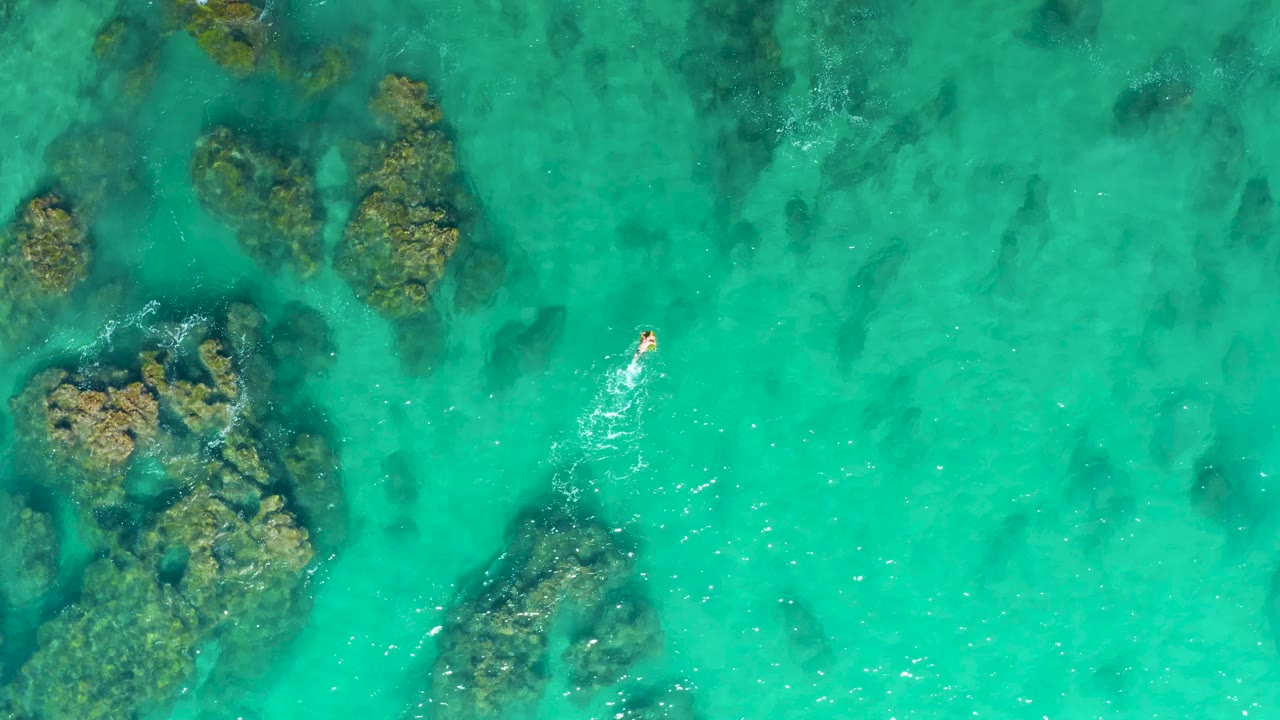 Aerial frame from Laguna Beach Los Cabos showing a small swimmer moving through clear turquoise water with large underwater rocks to the left.