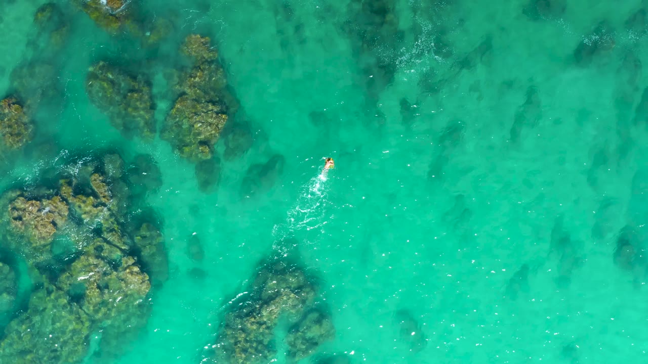 Aerial frame from Laguna Beach Los Cabos showing the swimmer near the upper right of the composition, with clustered underwater rocks below and left.