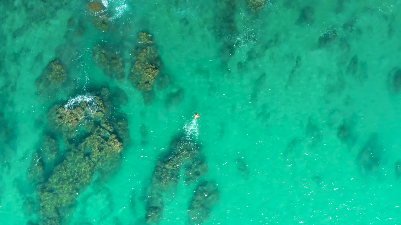At 0:02, the overhead frame shows Laguna Beach Los Cabos shallows with the person visible near the right side, a short white splash trail, and darker rocks beneath the water.