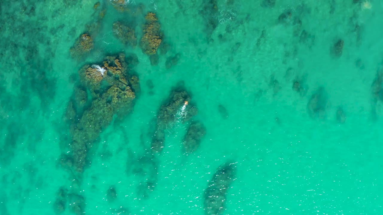 Aerial frame of Laguna Beach Los Cabos water with a larger rock formation left of center, a smaller submerged rock to the right, and no visible people.