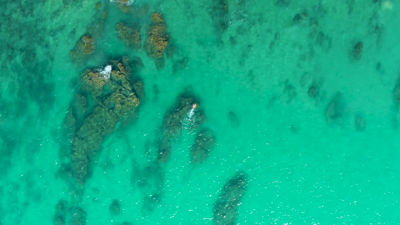 Top-down frame from Laguna Beach Los Cabos showing teal water, scattered brown rocks below the surface, faint foam near the upper rock, and no visible people.