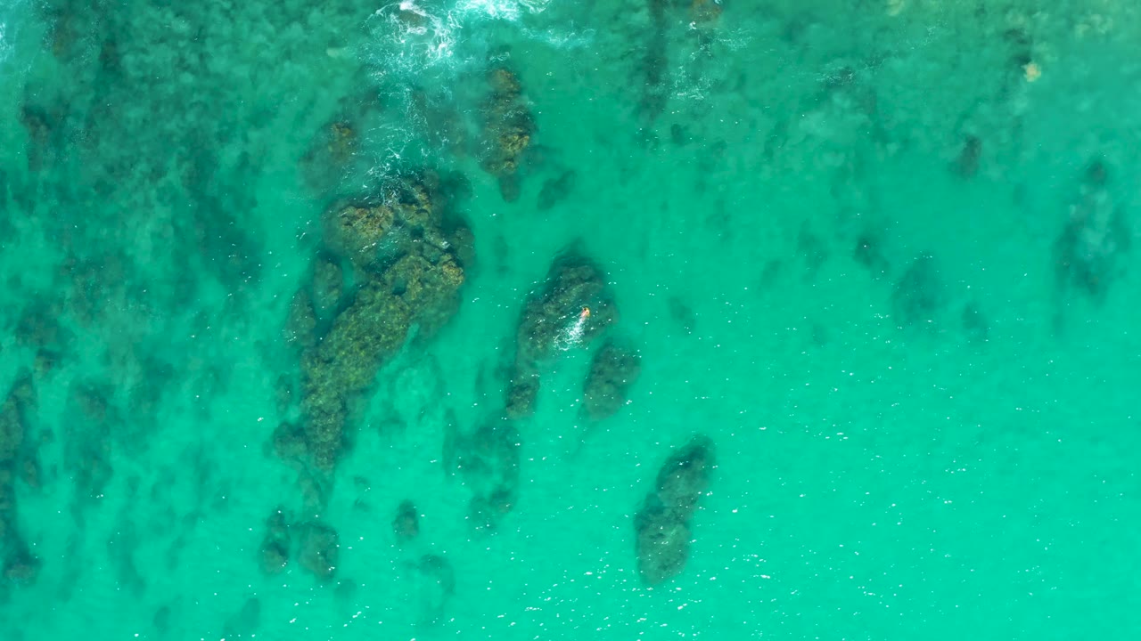 Overhead frame from Laguna Beach Los Cabos showing blue-green water, a central submerged rock formation, darker seabed patches, and no visible people.