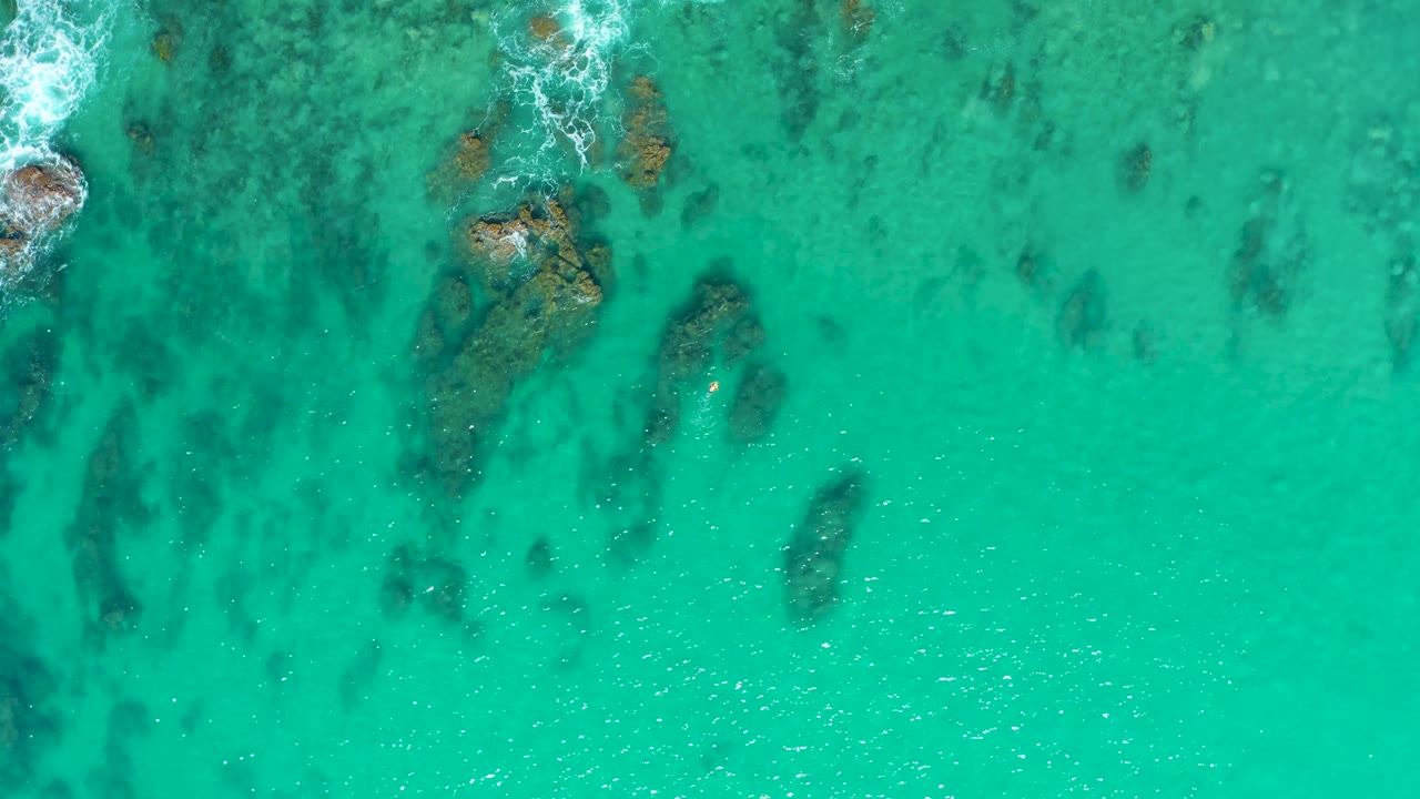 Aerial frame over Laguna Beach Los Cabos water, showing dark submerged rock shapes under clear turquoise sea and a small band of foam near the top, with no visible people.