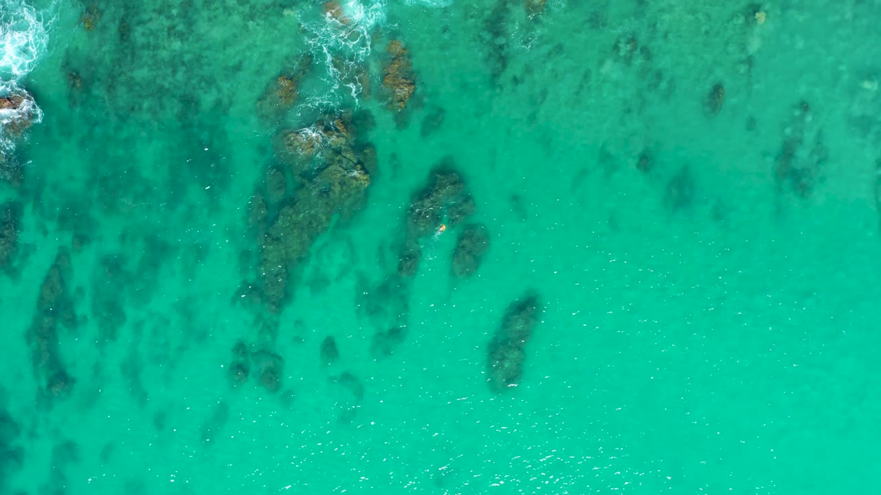 Top-down Laguna Beach Los Cabos sea view with muted dark rocks beneath turquoise water, light surface ripples, foam at the upper left edge, and no visible people.