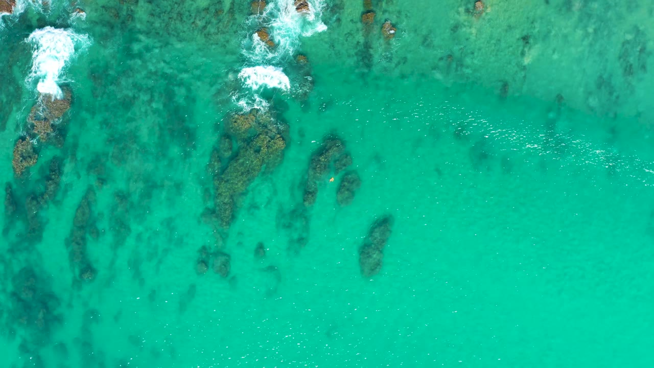 Aerial frame over Laguna Beach Los Cabos water with a larger rock cluster near the upper center, darker seabed shadows below it, small foam lines, and no visible people.