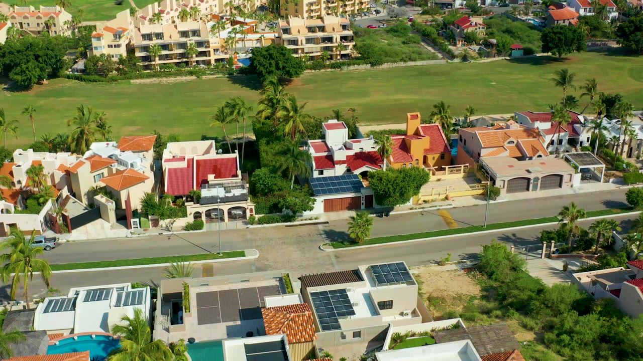 Aerial frame over San Jose Del Cabo homes with red roofs, palm trees, a road across the lower half, a green field behind the houses, and no visible people.