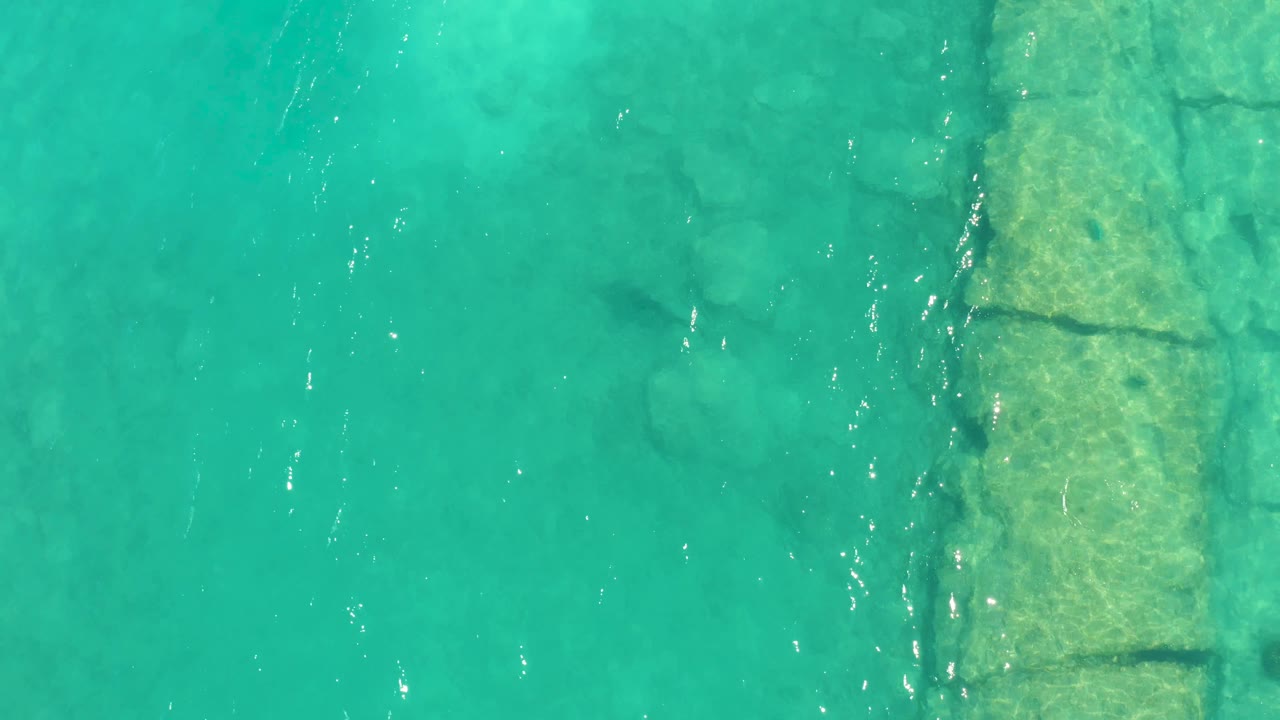 Clear turquoise water in Cabo Pulmo shows the stone edging nearer the center of frame, with light ripples moving across the shallow rocky bottom and no visible people.