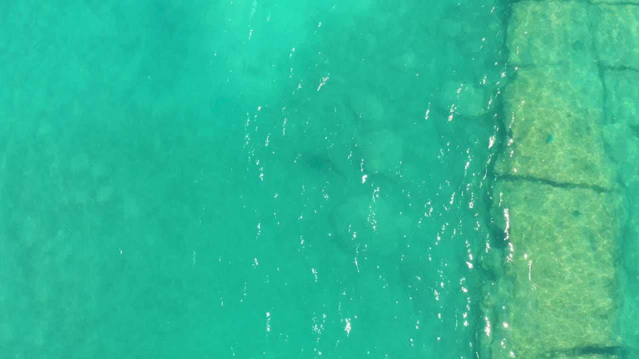Overhead Cabo Pulmo frame with turquoise water, a vertical submerged stone structure at left, faint rocks below the surface, and no visible people.