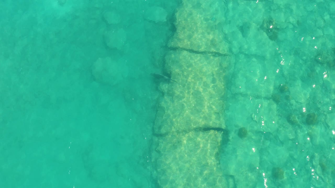 Clear Cabo Pulmo water shows the submerged stone structure slightly right of center, with ripples, pale rock texture, small fish shapes, and no visible people.